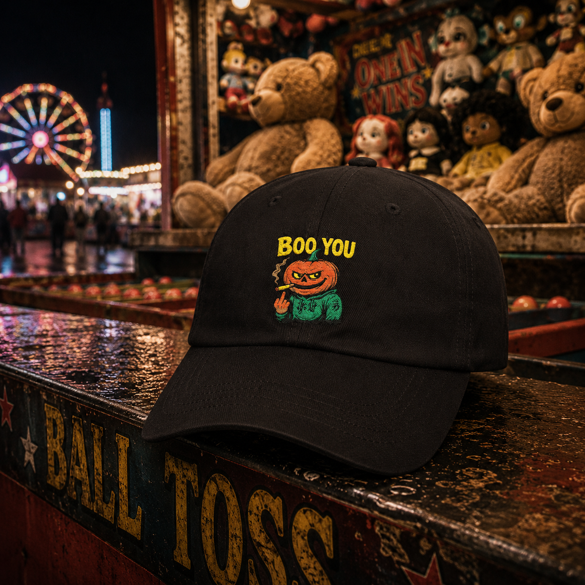 A black baseball cap with a colorful embroidered graphic of a smiling pumpkin character and the text 'BOO YOU' sits on a carnival ball toss table in front of a haunted house game booth filled with teddy bears at night.